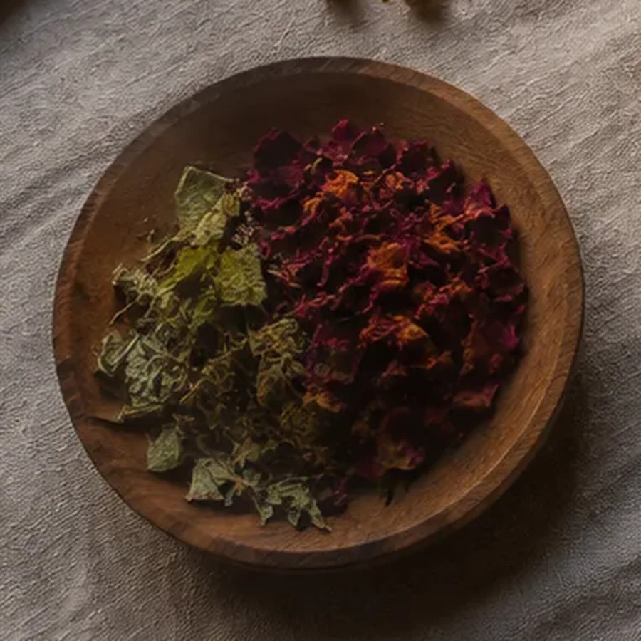Wooden dish of dried rose petals and herb leaves resting on a linen cloth